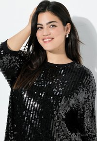 Schwarzer, mit Pailletten besetzter Pullover mit langen Ärmeln, runder Halsauschnitt und glatter Textur. Das Model fasst sich ins Haar und präsentiert Ohrringe.