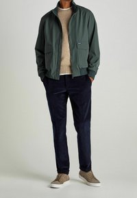 Giacca bomber verde con due tasche frontali, maglione in maglia beige, pantaloni in corduroy blu scuro e sneakers in suede marrone, abbinati insieme.