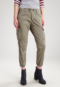 Olivgrüne Cargo-Hosen aus Baumwolle mit seitlichen Taschen, elastischen Bündchen und einer lockeren Passform. Getragen mit dunkelbraunen Knöchelstiefeln.