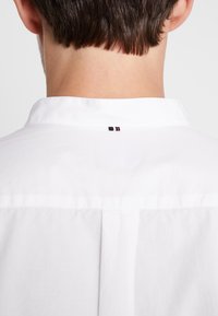 Biała koszula z kołnierzykiem o gładkiej fakturze, z małą metką z czerwono-białymi-niebieskimi paskami z tyłu na szyi.