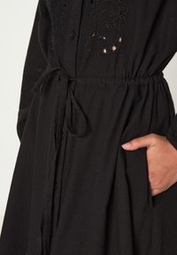 Robe noire à boutonnière avec des détails brodés et des découpes, dotée d'une taille à cordon et de poches latérales. Matière douce et texturée.