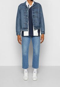 Jeansjacka med knäppning, blå färg, lager på en marinblå och vit pikétröja, tillsammans med ljusblå rakbenade jeans och vita sneakers.