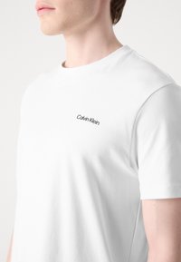 Jeune personne portant un t-shirt blanc uni Calvin Klein avec un petit logo sur le haut de la poitrine sur fond blanc.