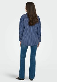 Chemise à manches longues rayée bleu et blanc, ayant une coupe décontractée avec un ourlet arrondi, associée à un jean évasé et des sandales.