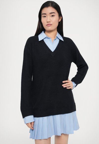 Fekete kötött pulóver V-nyakkivágással és hosszú ujjal, világoskék csíkos, galléros ing és pliszírozott szoknya fölött rétegezve.