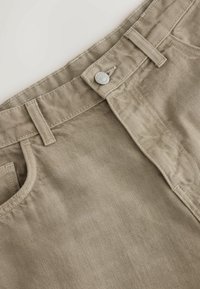 Gros plan sur la ceinture d'un jean beige en denim avec des passants de ceinture, une fermeture par bouton métallique et le bord supérieur de la poche avant visible.