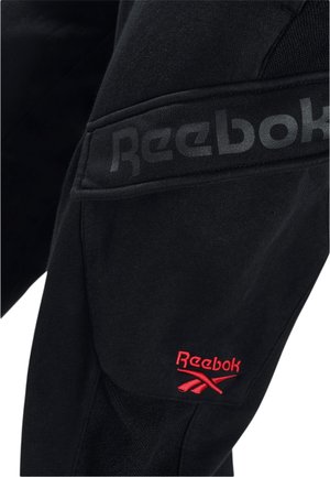 Schwarzer Stoff mit schwarzem Reebok-Logo oberhalb eines roten, gestickten Reebok-Logos und Vektorsymbols auf einer Tasche oder Kleidungsstück.