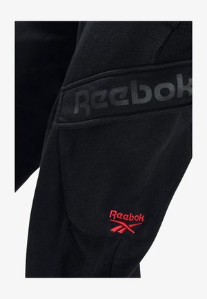 Schwarzer Stoff mit schwarzem Reebok-Logo oberhalb eines roten, gestickten Reebok-Logos und Vektorsymbols auf einer Tasche oder Kleidungsstück.