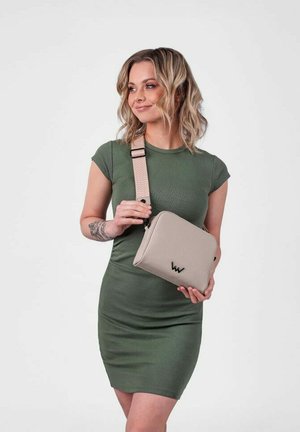 Beige crossbody taška s texturovaným popruhem, se zipovým uzávěrem a černým logem. Modelka má na sobě těsně přiléhající zelené šaty a stojí na světlém pozadí.