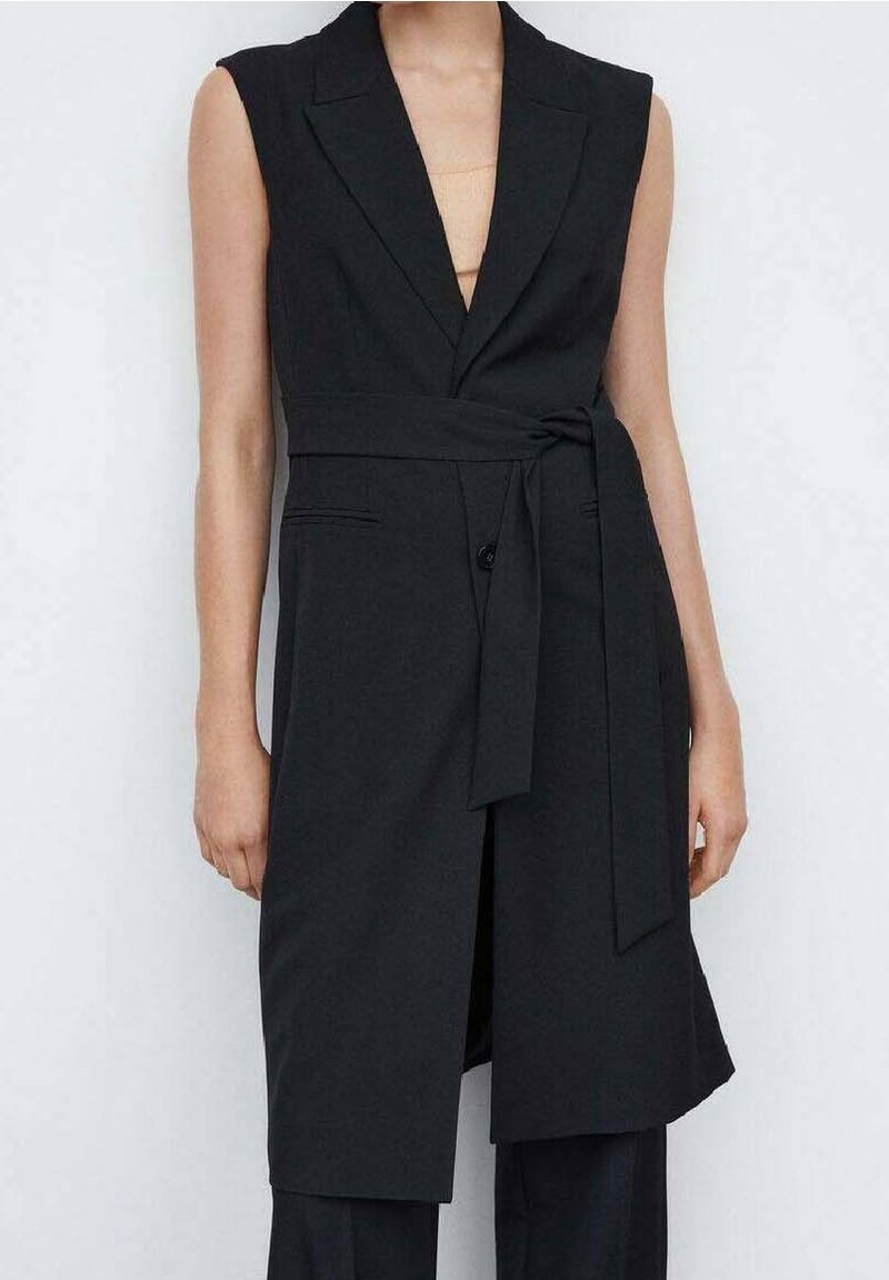 Blazer noir sans manches ceinturé avec revers, porté sur un haut beige et un pantalon noir, visible des épaules jusqu'à mi-cuisse.