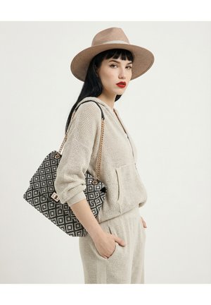 Femme en tenue en tricot beige et chapeau à large bord portant un sac à bandoulière noir et blanc à motif avec une bandoulière chaîne, debout la main dans la poche.