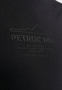 Tricou din bumbac negru cu text embossat. Textul include "PETROL IND" și detalii despre locație, împreună cu un grafic de munte în partea de sus.