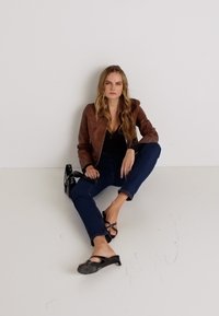 Rudas odinis bombonešio stilius, juoda palaidinė, tamsiai mėlynos džinsai ir juodi sandalai. Modelis sėdi ant grindų, laisva poza, minimalus fonas.