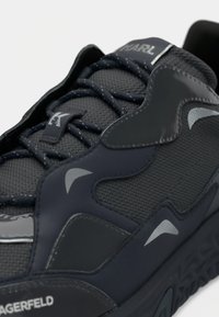 Crna sportska cipela koja kombinira teksturiranu mrežicu i glatki poliuretan. Ima srebrne naglaske i debeli gumeni potplat.