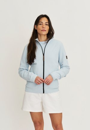 Vrouw met lang donker haar draagt een lichtblauwe trui met ritssluiting en witte short, staand tegen een effen achtergrond met gevlochten handen.