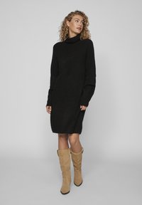Abito oversize nero in maglina con colletto alto e maniche lunghe, caratterizzato da una vestibilità comoda e spacchi laterali, abbinato a stivali beige al ginocchio.