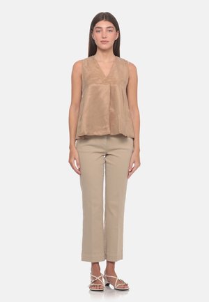 Donna con lunghi capelli castani che indossa un top smanicato in raso beige, pantaloni beige corti e sandali bianchi con tacco, in piedi davanti a uno sfondo semplice.