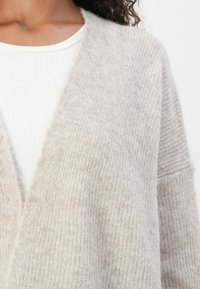 Light beige cardigan made of soft, textured fabric with a loose fit and open front. Layered over a white ribbed shirt.