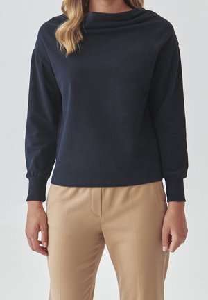 Frau trägt ein marineblaues Langarmoberteil mit umgeschlagenen Ärmeln und beige Hosen, stehend vor einem schlichten hellen Hintergrund.