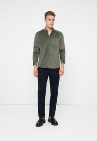 Muž v zelených kordových košilových šortkách, námořnicky modrých slim-fit nohaviciach a čiernych kožených mokasínach stojí na bielom pozadí.