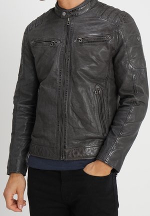 Mann trägt eine dunkelgraue Lederjacke mit Reißverschlüssen an Brust- und Seitentaschen, steht mit einer Hand in der Tasche und der anderen entspannt.