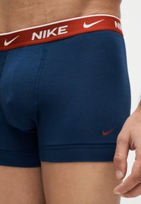 Κοντινό πλάνο ενός ατόμου που φοράει ναυτικό μπλε μποξεράκια Nike με κόκκινη μέση που φέρει λευκά λογότυπα Nike και κόκκινο λογότυπο Nike στη φάσα του ποδιού.