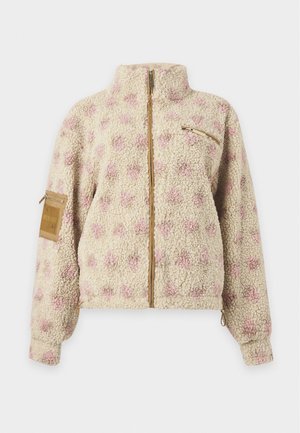 Femme portant une veste zippée beige texturée avec des pois roses, une longue jupe blanche avec un bord en dentelle et des chaussures marron à lacets, debout avec une main dans la poche.