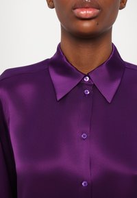 Lilla satin skjorte med klassisk krave, fem knapper og glat tekstur. Stoffet har en let glans, der fremhæver den levende farve.