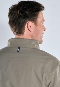 Olive green jacket with a stand collar, featuring a logo tag at the back and smooth fabric texture. Minimalist design and tailored fit.