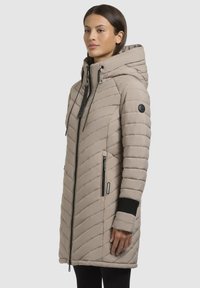 Abrigo de invierno acolchado de color beige con capucha, que presenta patrones de acolchado diagonales, bolsillos con cremallera, detalles en negro y puños de manga acanalados.