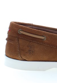 Ruskea mokkanahkainen venekenkä valkoisella kumipohjalla, jossa on koristeellinen ommel ja pieni logoyksityiskohta kantapään lähellä. Ruskeat yksityiskohdat korostavat tekstuuria.