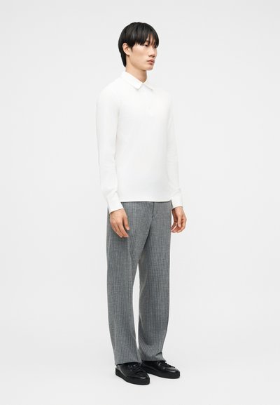 Άνδρας που στέκεται, φοράει εφαρμοστή λευκή μακρυμάνικη μπλούζα, γκρι παντελόνι με λεπτές ρίγες και μαύρα αθλητικά παπούτσια, μπροστά σε απλό λευκό φόντο.