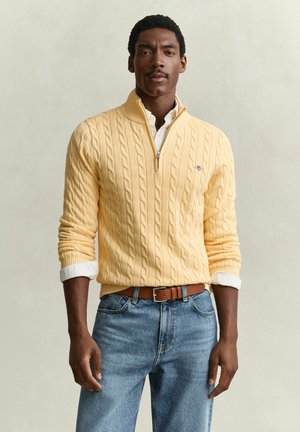 Homme portant un pull jaune à demi-fermeture éclair en maille torsadée par-dessus une chemise blanche, jean bleu et ceinture marron, debout devant un fond uni.