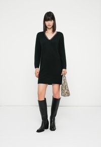 Schwarzes Strickkleid mit V-Ausschnitt und langen Ärmeln, verziert mit einer Spitzenborte. Kombiniert mit kniehohen schwarzen Stiefeln und einer Handtasche im Schlangenlederdruck.