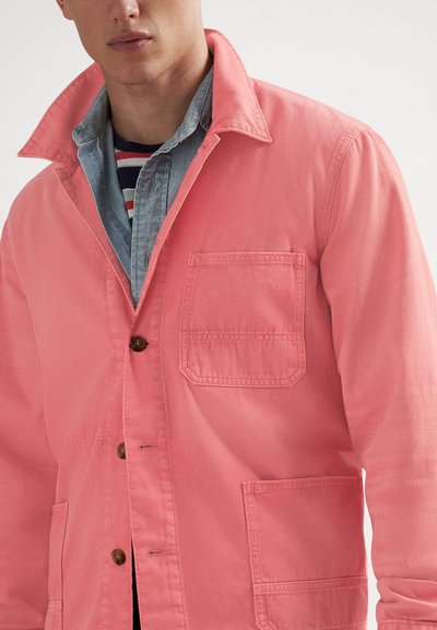 Vīrietis, kurš valkā vairākas drēbes — virsū rozā poga āda jaka, zem tās gaiša džinsa krekls un svītrains t-krekls.