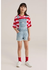 Mladá dívka stojí s rukama v kapsách, nosí červené a růžové pruhované tričko, světle modré denimové krátké lacláče, červené ponožky a bílé tenisky.