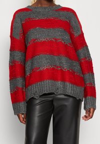 Person som bär en löst sittande röd och grårandig stickad tröja med slitna detaljer och svarta läderbyxor, visad från axlarna till höfterna.