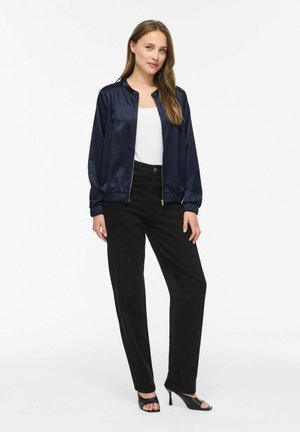Femme debout portant une veste bomber en satin bleu marine, un haut blanc, un pantalon noir et des talons ouverts noirs sur un fond blanc uni.
