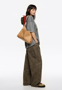 Borsa a tracolla in pelle color cuoio con un design morbido. Modello in una maglietta a maniche corte grigia con grafica e pantaloni larghi a motivo verde oliva.