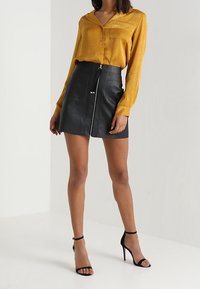 Blouse en satin moutarde à manches longues et poche, associée à une mini-jupe en simili cuir noire avec fermeture éclair sur le devant et quincaillerie en argent.