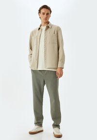 Camisa beige de botones sobre una camiseta blanca, combinada con pantalones de chándal verde oliva y zapatillas blancas con detalles en marrón. Diseño sencillo, corte holgado.