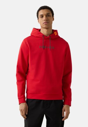 Jonge man met een rode capuchontrui met de tekst "FIRE + ICE", staand tegen een effen lichte achtergrond.