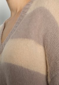 Närbild av en person som bär en mjuk, randig stickad tröja i beige och taupe med V-hals.