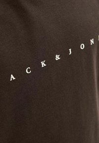 Ruda medvilninė marškinėliai su iškiliais baltais rašmenimis, kurie sako "ACK & JON", prieš lygaus tekstūruoto fono.