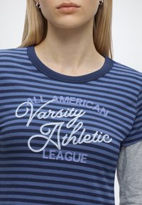 Жена, облечена в синя райета риза с текст "All American Varsity Athletic League" в бял и светлосин шрифт.