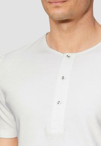 Witte T-shirt met korte mouwen en ronde halslijn, voorzien van een placket met vier knopen. Zachte textuur met subtiele stikseldetails.