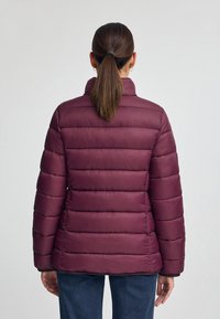 Bourgogne puffjacka med horisontellt kviltat mönster, hög krage och elastiska ärmslut, tillsammans med blå denimjeans.