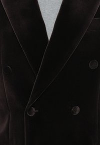 Blazer de veludo preto de dupla abertura com lapelas largas e botões cobertos pelo mesmo tecido, sobre um top de malha cinza claro.