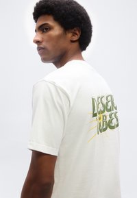 Νέος άντρας με κοντά σγουρά μαλλιά, φοράει λευκό μπλουζάκι με το κείμενο «DESERT VIBES» και γραφικό κίτρινο ήλιο στην πλάτη, κοιτάζοντας πλάγια.