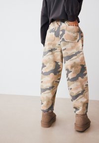 Pantalon ample à motif camouflage dans des tons beige et gris, avec une taille élastique et une poche arrière, associé à des chaussures basses marron.